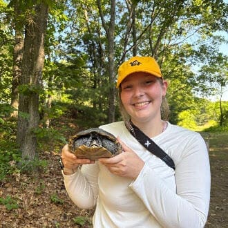 Amethyst Clark, the article’s author, smiling in a yellow cap and holding a diamondback terrapin on a wooded trail
